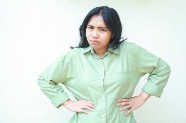 Huysuz Asyalı kadın elleri belinde kameraya bakıyor yeşil günlük kıyafetler giyiyor çılgın öfkeli bir ifade sergiliyor, beyaz arka planda izole edilmiş.