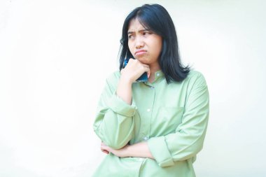 Asyalı üzgün bir kadının portresi. Kollarını kavuşturmuş, çenesinde yeşil tişört, elinde akıllı telefon, boş alana bakıyor. Endişeli düşünce sorunu, izole beyaz arka planda duruyor.