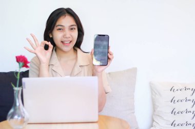 Güzel Asyalı bir kadın rahat bir kanepeye oturup akıllı telefon beyaz ekranı gösterir ve ok işaretleri verir. Ahşap masaya dizüstü bilgisayarı olan günlük kıyafetler giyer, uygulamaları tanıtım konsepti uygular.