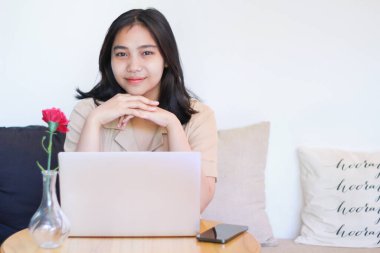 Mutlu Asyalı kadın, dizüstü bilgisayar ve akıllı telefon kullanarak koltukta oturuyor. Gülümseyen kameraya bakıyor.