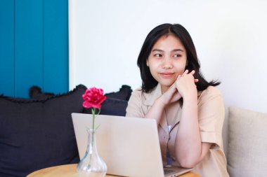 Mutlu Asyalı kadın dizüstü bilgisayar ve akıllı telefon kullanarak koltukta oturuyor. Gülümseyerek başka tarafa bakıyor. Günlük kıyafetler giyiyor.