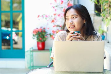 Mutlu Asyalı kadın kafede dizüstü bilgisayarla çalışırken kahve içer açık havada gündelik kıyafetler giyer, şık giyinir, uzaktan kumandalı kadın güler yüzlü başka yere bakar.