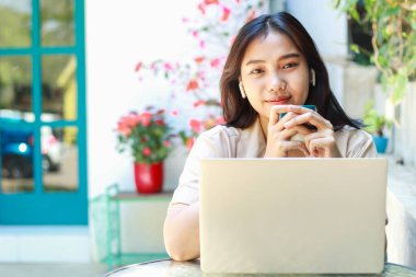 Mutlu Asyalı kadın kafede dizüstü bilgisayarla çalışırken kahve içer açık havada gündelik giysiler giyer, uzaktan kumandalı kadın kameraya bakar.