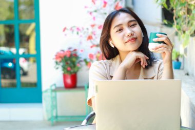 Mutlu Asyalı kadın kafede dizüstü bilgisayarla çalışırken kahve içer açık havada gündelik giysiler giyer, uzaktan kumandalı kadın kameraya bakar.