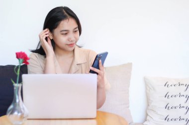 Mutlu Asyalı kadın rahat bir koltukta oturuyor. Akıllı telefonu ahşap masada dizüstü bilgisayarı var. Şık kıyafetler giyiyor.