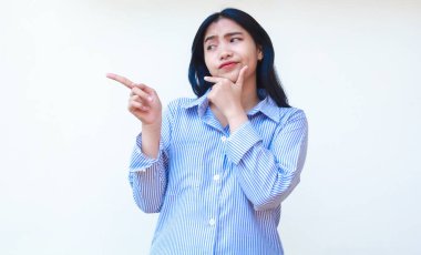 Kafası karışmış Asyalı kadın, sıradan çizgili tişört giyip boş bir yeri işaret ederek izole edilmiş beyaz arka planda duran seçenekleri düşünüyor.