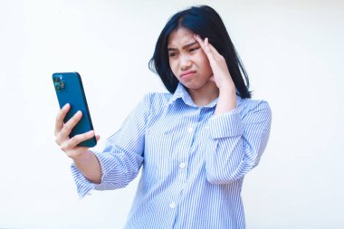 Akıllı telefonu kullanan düşünceli Asyalı kadın kafasında mavi çizgili gündelik gömleği olan yalnız beyaz arka planda duran, kafasını çevirmiş.