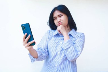Akıllı telefon kullanan düşünceli Asyalı kadın çene düşünme problemi yaşıyor mavi çizgili günlük gömlek giyiyor izole beyaz arka planda dikiliyor, başka tarafa bak.