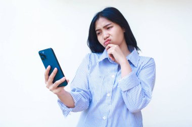 Akıllı telefon kullanan düşünceli Asyalı kadın çene düşünme problemi yaşıyor mavi çizgili günlük gömlek giyiyor izole beyaz arka planda dikiliyor, başka tarafa bak.