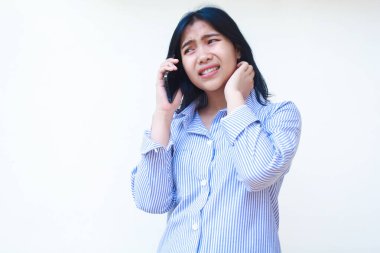 Asyalı düşünceli bir kadın boynunda akıllı telefon kullanıyor mavi çizgili bir gömlek giyiyor ve beyaz arka planda dikiliyor, başka tarafa bakıyor.
