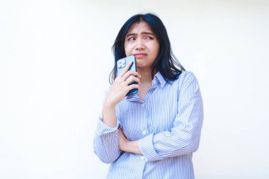 Akıllı telefon kullanan düşünceli Asyalı kadın çene düşünme problemi yaşıyor mavi çizgili günlük gömlek giyiyor izole beyaz arka planda dikiliyor, başka tarafa bak.