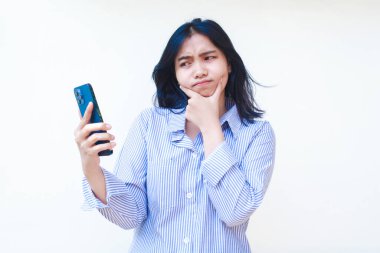 Akıllı telefon kullanan düşünceli Asyalı kadın çene düşünme problemi yaşıyor mavi çizgili günlük gömlek giyiyor izole beyaz arka planda dikiliyor, başka tarafa bak.