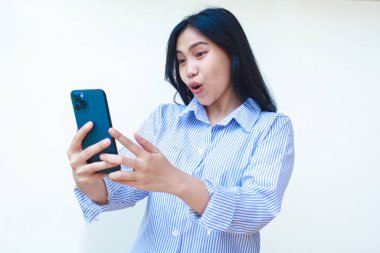 Cep telefonu kullanan mutlu Asyalı kadın beyaz arka planda izole edilmiş çizgili gömlek giyerek sosyal medyada sörf yaparken şok oldu.
