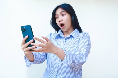 Şok olmuş Asyalı kadın beyaz arka planda izole edilmiş çizgili gömlek giymiş ağzı açık bir şekilde akıllı telefon kullanıyor.