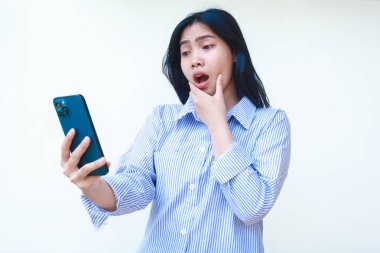Şok olmuş Asyalı kadın beyaz arka planda izole edilmiş çizgili gömlek giymiş ağzı açık bir şekilde akıllı telefon kullanıyor.