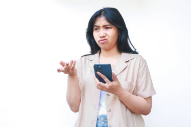 Kafası karışmış Asyalı bir kadın avuçlarını kaldırmış akıllı telefon kullanıyor. Belirsizlik ifadesi var. Beyaz arka planda izole edilmiş günlük kıyafetler giyiyor.