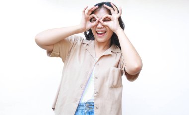 Tatmin olmuş Asyalı bir kadın, beyaz arka planda izole edilmiş günlük kahverengi kıyafetler giyerek heyecan verici işaretler gösteriyor.