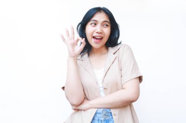 Tatmin olmuş Asyalı bir kadın, beyaz arka planda izole edilmiş günlük kahverengi kıyafetler giyerek heyecan verici işaretler gösteriyor.