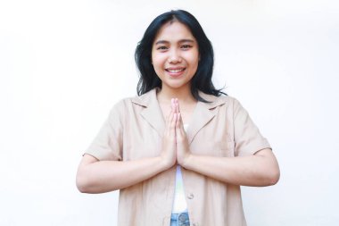 Kameraya gülümseyen mutlu Asyalı kadın geleneksel selamlama jestiyle beyaz arka planda izole edilmiş kahverengi elbiseler giyiyor.