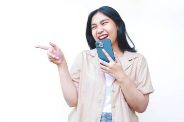 Heyecanlı Asyalı kadın elinde akıllı telefon, beyaz arka planda izole edilmiş kahverengi günlük giysiler giyiyor. Ürün satış promosyonu kavramı.