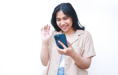 Heyecanlı Asyalı kadın beyaz arka planda izole edilmiş günlük kahverengi kıyafetler giyerek video görüşmesinde konuşurken akıllı telefon ve el sallıyor.