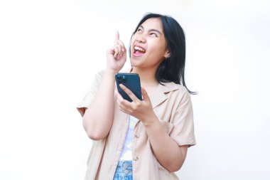 Heyecanlı Asyalı kadın elinde akıllı telefon, beyaz arka planda izole edilmiş kahverengi günlük giysiler giyiyor. Ürün satış promosyonu kavramı.