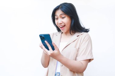 Heyecanlı Asyalı kadın beyaz arka planda izole edilmiş günlük kahverengi kıyafetler giyerek akıllı telefon kullanırken şaşırdı.