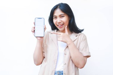 Heyecanlı Asyalı kadın, beyaz ekranı işaret ederek jest yapıyor beyaz arka planda izole edilmiş kahverengi günlük kıyafetler giyiyor.