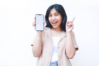 Heyecanlı Asyalı kadın, beyaz ekranı işaret ederek jest yapıyor beyaz arka planda izole edilmiş kahverengi günlük kıyafetler giyiyor.