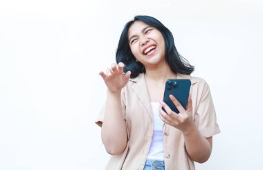 Zeki telefon kullanan mutlu Asyalı kadın beyaz arka planda izole edilmiş günlük kahverengi kıyafetler giyen, akıllı telefonu kaldırarak gülüyor.