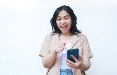 Mutlu Asyalı kadın gülüyor, akıllı telefon beyaz arka planda izole edilmiş günlük kahverengi kıyafetler giyerek sosyal medyaya kayıyor.
