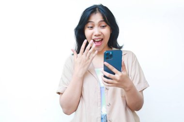 Akıllı telefon tutan Asyalı kadın beyaz arka planda izole edilmiş günlük kıyafetler giyerken çok şaşırmış görünüyor.