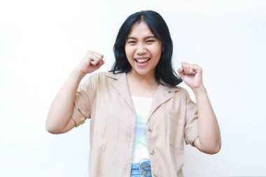 Mutlu Asyalı kadın zaferi, beyaz arka planda izole edilmiş günlük kahverengi giysiler içinde yumruğunu kaldırarak kutluyor.