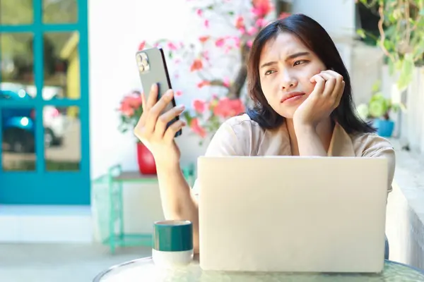 confused asian woman holding smartphone with laptop on table sitting in outdoors cafe working space wearing casual clothes