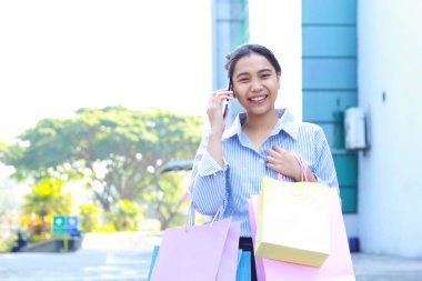 Neşeli Asyalı kadın alışveriş çantası taşıyor ve dışarıda yürürken cep telefonuyla konuşuyor.