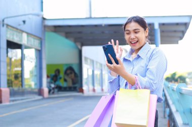 Neşeli Asyalı kadın alışveriş çantası taşıyor ve elinde akıllı telefonuyla alışveriş merkezinde yürürken selfie çekiyor.