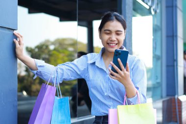 Neşeli Asyalı kadın alışveriş çantası taşıyor ve elinde akıllı telefonuyla alışveriş merkezinde yürürken selfie çekiyor.