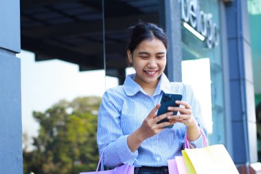 Neşeli Asyalı kadın alışveriş çantası taşıyor ve elinde akıllı telefonuyla alışveriş merkezinde yürürken selfie çekiyor.