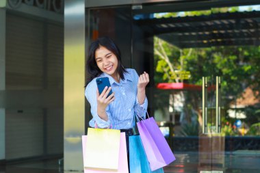 Alışveriş çantası taşıyan mutlu Asyalı kadın ve elinde akıllı telefon ve yumruklanmış zafer jesti dışarıda alışveriş merkezinin etrafında yürüyor.