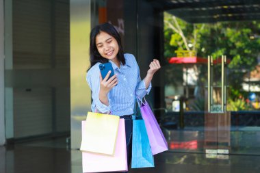 Alışveriş çantası taşıyan mutlu Asyalı kadın ve elinde akıllı telefon ve yumruklanmış zafer jesti dışarıda alışveriş merkezinin etrafında yürüyor.