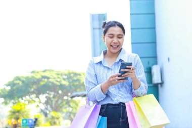Alışveriş çantası ve akıllı telefon taşıyan gülen Asyalı kadın şehir merkezinin etrafında dolanıyor.