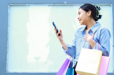 Asyalı bir kadının alışveriş çantası taşımasına ve şehir merkezinin yakınındaki boş bir billborda arka planına sahip açık alanda akıllı telefon kullanmasına şaşırdım.
