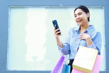 Asyalı bir kadının alışveriş çantası taşımasına ve şehir merkezinin yakınındaki boş bir billborda arka planına sahip açık alanda akıllı telefon kullanmasına şaşırdım.
