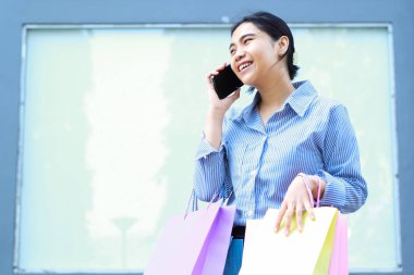 Elinde alışveriş çantası olan gülümseyen Asyalı kadın. Akıllı telefonuyla konuşuyor. Açık havada duruyor.