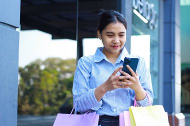 Alışveriş torbaları tutan ve akıllı telefon kullanan gülümseyen Asyalı kadın alışveriş merkezinin etrafında yürürken tatmin oluyor.