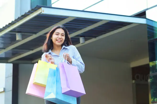 happy beautiful asian woman holding shopping bag while walking in outdoors around mall building with excitement