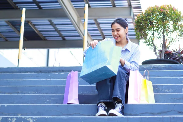 low angle view of happy beautiful asian woman checking her shopping bag while sitting in outdoors staircase around mall building with excitement, full body glamour female