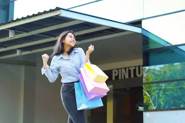 excited asian woman holding shopping bag celebrate victory with raised hands show yes gesture while walking in outdoors around mall building