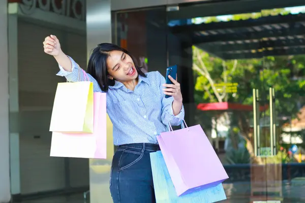 happy asian woman carrying shopping bag and holding smartphone with raised fist celebrating victory gesture walking in outdoors around mall building