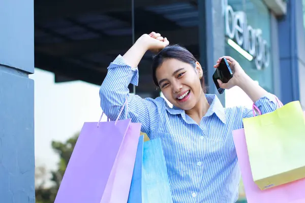happy asian woman carrying shopping bag and holding smartphone with raised fist celebrating victory gesture walking in outdoors around mall building